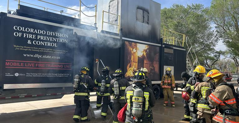 A group of firefighters undergoing realistic structural fire suppression training. They are positioned at the entrance of the Colorado Division of Fire Prevention and Control’s Mobile Live Fire Training Unit, which features a large graphic of a fire on its side and scorched upper panels from heat exposure.